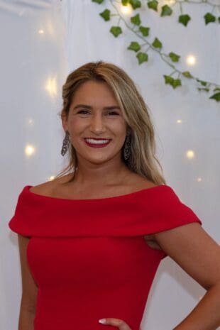 W&J junior Alyssa Pollice stands in front of while backdrop in red dress and smiles.