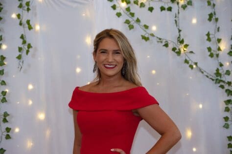 W&J junior Alyssa Pollice stands in front of while backdrop in red dress and smiles.