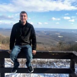 Erik Blasic sits on a fence in front of a mountainous backdrop.