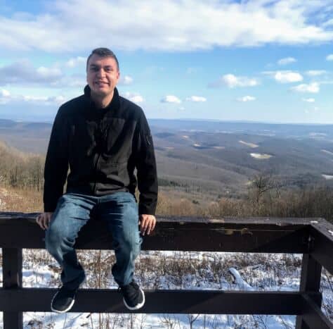 Erik Blasic sits on a fence in front of a mountainous backdrop.