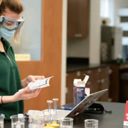 A student wearing goggles and a mask stands by a desk in a science lab with beakers in front of her.