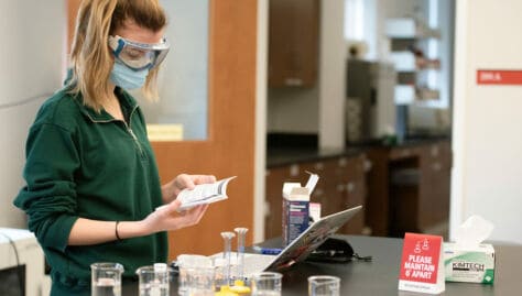 A student wearing goggles and a mask stands by a desk in a science lab with beakers in front of her.