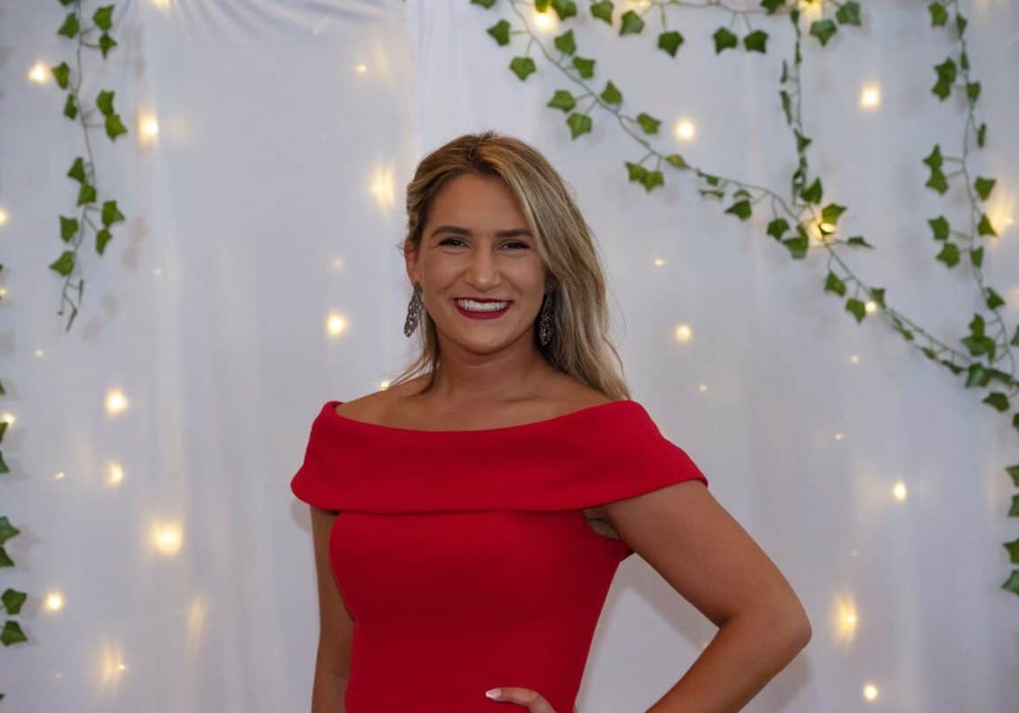 W&J junior Alyssa Pollice stands in front of while backdrop in red dress and smiles.