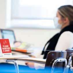 A student sits at her desk on her laptop in the background. In the foreground is a sign that reads "please maintain 6 feet" resting on a desk that is roped off.