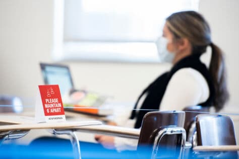 A student sits at her desk on her laptop in the background. In the foreground is a sign that reads "please maintain 6 feet" resting on a desk that is roped off.