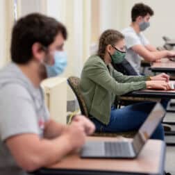 Students sit in their class on laptops at desks that are socially distanced. Students are wearing masks.