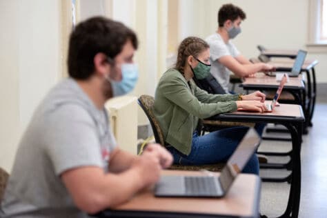 Students sit in their class on laptops at desks that are socially distanced. Students are wearing masks.